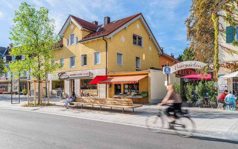 Eine neu gestalteten Straße in einer Kleinstadt mit frisch gepflanzten Bäumen, breiten Fußgängerwegen, großer Sitzbank und Radfahrerin im Vordergrund.