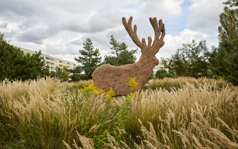 Hochgewachsene Gräser, in der Mitte eine Hirsch-Skulptur, dahinter Plattenbauten